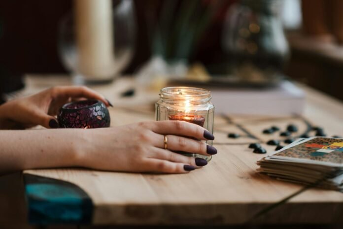 Hands with dark nails holding a lit candle, surrounded by tarot cards, creating a mystical and warm ambiance.